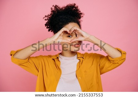 Lovely candid pleasant african american girl, makes heart gesture with hands near eye, demonstrates love sign, stands on isolated pink background, looking at camera though love gesture, smile friendly