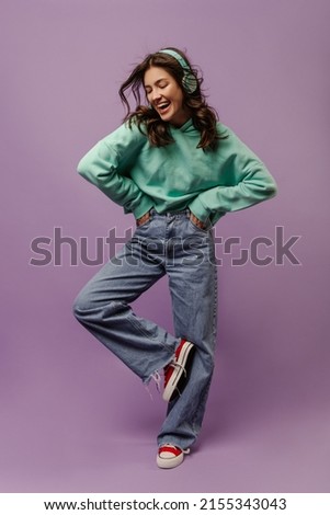 Full length happy young caucasian woman spends leisure time listening to favorite song through headphones in studio. Brunette wear casual clothes keeps hands in pockets and looks down at her feet. Royalty-Free Stock Photo #2155343043