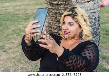pretty plus size caucasian woman with blonde hair sitting outdoors in the park, leaning against a palm tree, taking a selfie with her phone