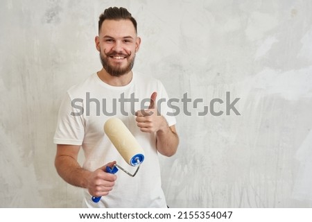 a young man with a mustache in a white t-shirt holds a wall paint roller in his hands and raises his thumb up, meaning that everything is cool