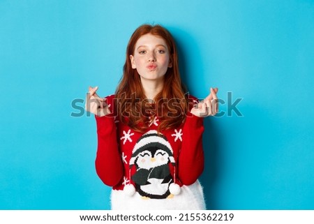 Winter holidays and Christmas Eve concept. Lovely redhead girl in xmas sweater, showing heart sign and pucker lips for kiss, standing over blue background