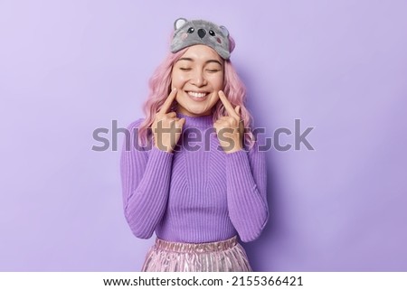Look at my toothy smile. Pleased pink haired beautiful young Asian woman points at her teeth keeps eyes closed feels very happy wears sleepmask and turtleneck isolated over purple background