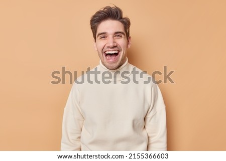 Waist up shot of overjoyed handsome man laughs happily expresses sincere emotions hears something funny dressed in casual white jumper isolated over brown background. People and happiness concept Royalty-Free Stock Photo #2155366603