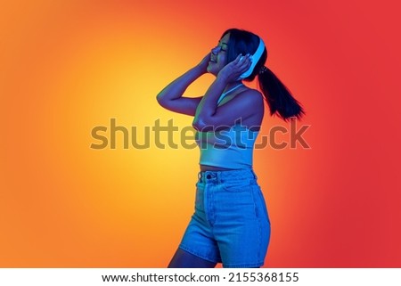 Young cheerful woman listening to music in headphones isolated over gradient yellow orange background in neon light. Concept of beauty, youth, facial expression, emotions, lifestyle. Copy space for ad