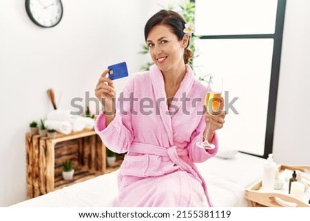 Middle age hispanic woman toasting with champagne holding credit card at beauty center