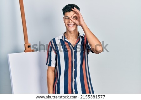Young hispanic man standing close to empty canvas doing ok gesture with hand smiling, eye looking through fingers with happy face. 
