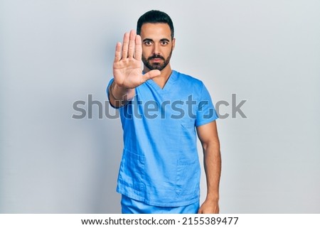 Handsome hispanic man with beard wearing blue male nurse uniform doing stop sing with palm of the hand. warning expression with negative and serious gesture on the face. 