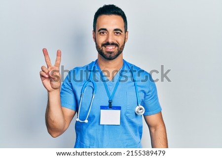 Handsome hispanic man with beard wearing doctor uniform smiling with happy face winking at the camera doing victory sign. number two. 