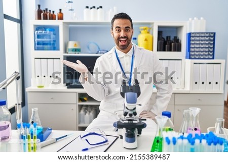Young hispanic man with beard working at scientist laboratory smiling cheerful presenting and pointing with palm of hand looking at the camera. 