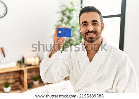 Young hispanic man wearing bathrobe holding credit card at beauty center