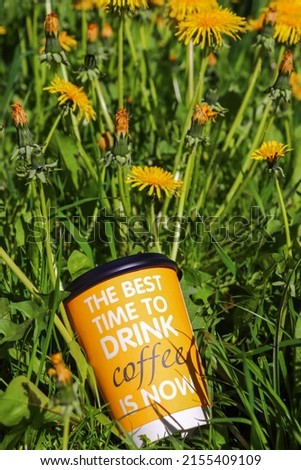 A cardboard glass for drinks in yellow color and text on it. In background there are grass and dandelions.
