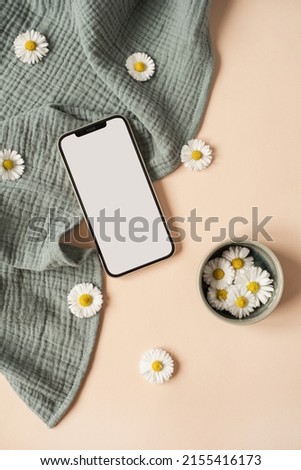 Flatlay of blank screen mobile phone, chamomile flower buds, aquamarine muslin cloth on neutral beige background. Aesthetic social media, blog template with mockup space. Flat lay, top view.