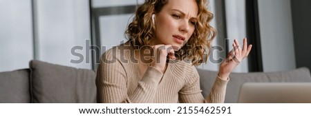 Young woman in earphones gesturing and working with laptop while sitting on couch at home