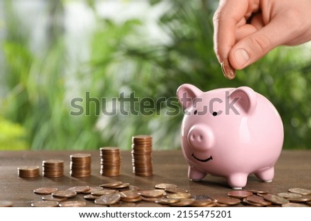 Woman putting coin into piggy bank at wooden table against blurred green background, closeup. Space for text