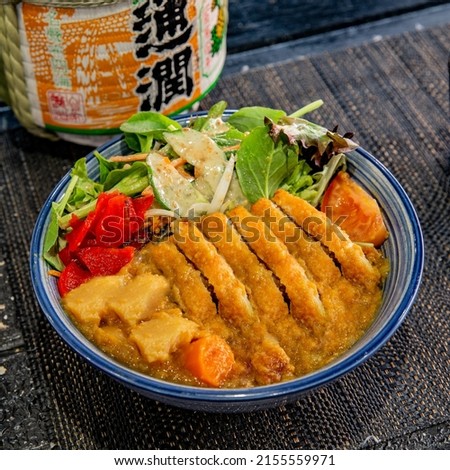 Fried chicken schnitzel curry rice with salad in the traditional Japanese ramen restaurant, with a black plate on a black mat and Japanese fonts sake barrel translation: Rich