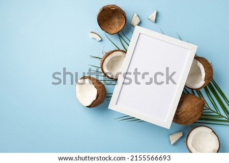 Summer holidays concept. Top view photo of white photo frame over cracked coconuts and palm leaves on isolated light blue background with empty space