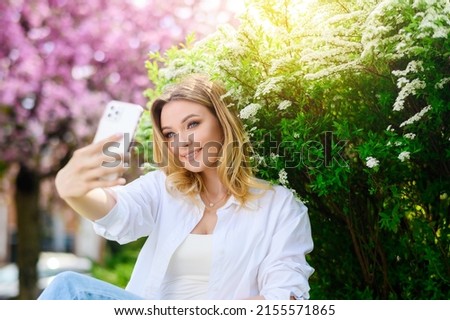 Beautiful blonde hair girl in a white shirt holds a phone and takes a selfie in the park. Sunshine