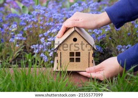 Hand holding a wooden house model against a flower bed outdoors. Springtime house decoration concept.