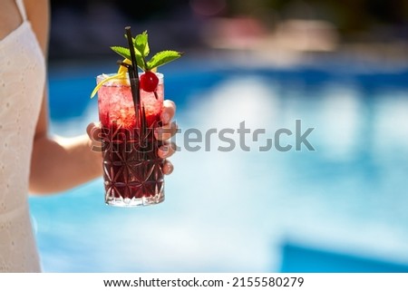 Close-up of woman hand cocktail glass decorated with mint, cherry and lemon. Female holding a drink near swimming pool with blue water. Girl chilling with beverage in tropical sun. Vacation concept