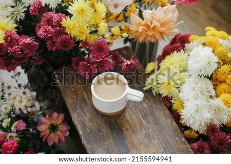 Warm cup of coffee and beautiful autumn flowers on rustic wooden background. Good morning. Stylish mug with cappuccino among colorful asters and dahlias composition. Moody image