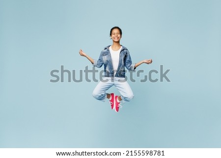 Full body little kid teen girl of African American ethnicity 12-13 years old in denim jacket jump high spread hands in yoga om gesture relax meditate isolated on pastel plain light blue background.