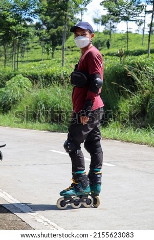 Selective focus of an Asian man in red who was learning to use roller skates on a concrete road with a panoramic view of a fresh green tea plantation. Commemorating World Skate Day