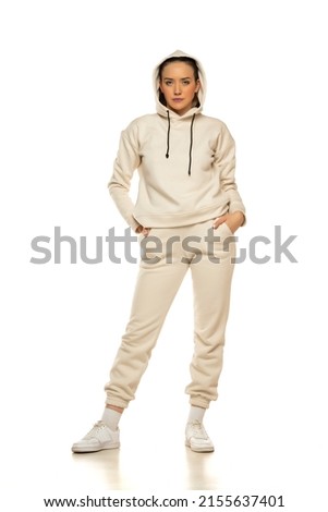 Front view of a young woman in a beige tracksuit with hood posing to a white background in the studio.