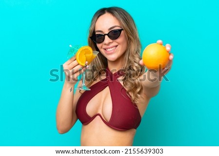 Young caucasian woman isolated on blue background in swimsuit and holding a cocktail