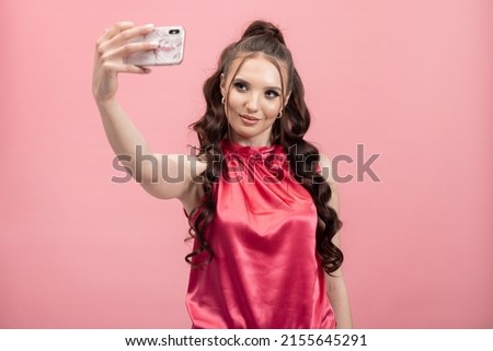 Portrait of a cheerful, positive, smiling woman holding a smartphone taking a selfie against a pink background.