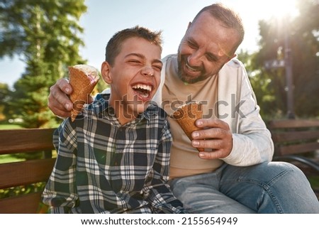 Cheerful caucasian father and teenage son with cerebral palsy eat ice cream and joke on bench in sunny park. Family relationship and enjoy time together. Disability care, treatment and rehabilitation Royalty-Free Stock Photo #2155654949