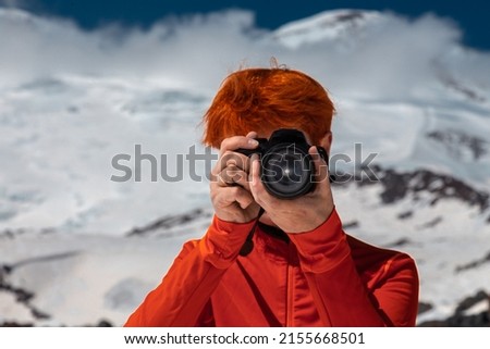 Red-haired girl with a face hidden behind a camera aimed directly at the frame against the backdrop of mountains