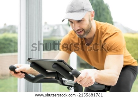 Hispanic young man with a stubble wearing a grey cap and orange top exercising on a spinning bike in a conservatory while following an online fitness workout on his tablet resting on the handlebar Royalty-Free Stock Photo #2155681761