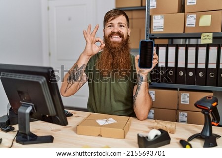 Redhead man with long beard working at small business holding smartphone doing ok sign with fingers, smiling friendly gesturing excellent symbol 