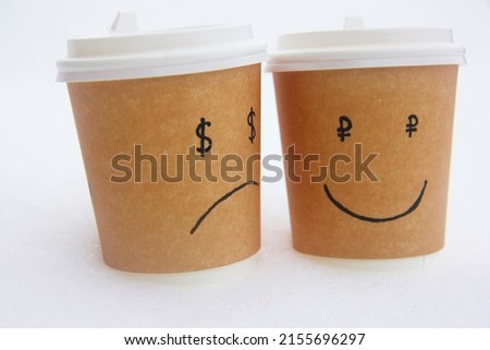 Coffee cups, on which a smile is drawn and in the eyes a sign of the ruble and chagrin and a dollar sign in the eyes.  The cups are on the snow.