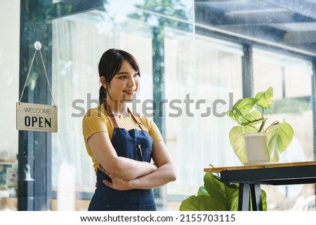Business owner happy beautiful young asian woman in apron, we're open sign on front door smiling welcoming clients to new cafe, People and start-up concept.