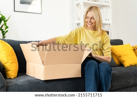 Happy senior mature adult woman is looking at her new parcels, sitting on the couch, and unpacking. Smiling lady opening online store order in the postal delivery shipping box Royalty-Free Stock Photo #2155711715
