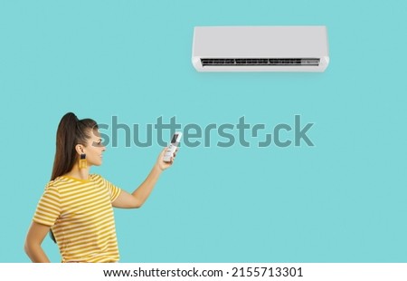 Happy woman for comfort during summer heat uses modern air conditioner, isolated on turquoise background. Smiling millennial woman in yellow T-shirt uses remote control to turn on air conditioner. Royalty-Free Stock Photo #2155713301