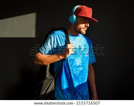 Man skateboarder with covered skateboard in morning sunlight. Listens music in blue headphones.