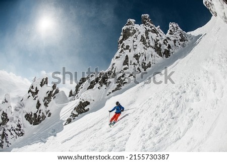 Skier dressed in bright sportswear with fast sliding down snow-covered slopes on skis. Another skier with a camera takes a photo on the background.