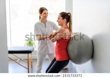 A Physiotherapist helping woman do wall squats with fit ball. Young female patient doing back exercise using fitball in physio room of modern clinic Royalty-Free Stock Photo #2155733373