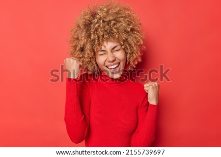 Positive curly haired young woman makes yes gesture celebrates success feels overjoyed and triumphant wears casual turtleneck shakes fists exclaims from joy isolated over vivid red background