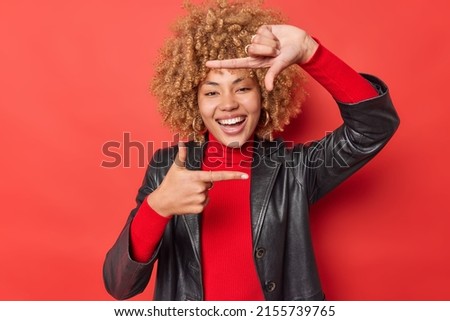 Positive woman makes frame gesture captures moment takes measurement pictures something smiles happily wears casual turtleneck and leather jacket being in good mood isolated over red background.