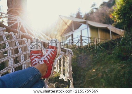 Cropped shot of female feet lying on the net hammock on the country house yard. Legs of a woman wearing blue jeans and red white snickers, relaxing on a beautiful day. Close up, copy space, background