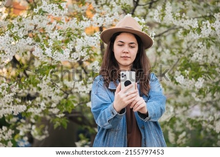 A smiling girl in a hat takes a selfie against the backdrop of a flowering tree. Spring background. Spring picture. Asian girl taking a selfie