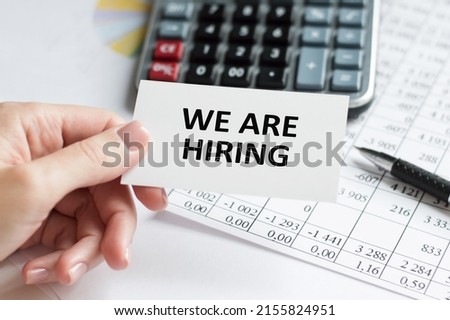 the text we are hiring on a business card in a person's hand against a background of reports, pens and calculator