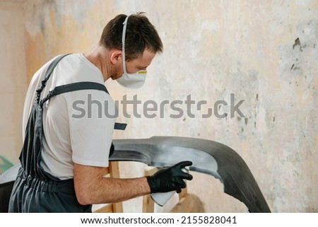 Young man painting a car bumper with spray can. Overhaul cars plastic bumper cover at auto repair shop. Worker using protective gear while working to keep from inhaling harmful fumes. Selective focus. Royalty-Free Stock Photo #2155828041