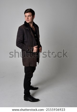 Full length portrait of  handsome brunette male model wearing black leather coat and elegant vest. Standing Pose with gestural hands reaching out,  isolated on studio background.