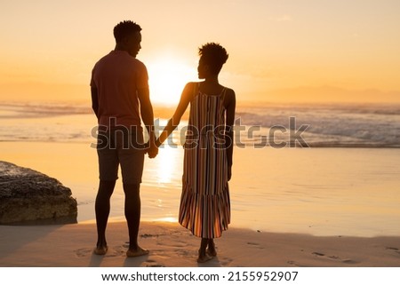 Rear view of african american young couple holding hands and standing on beach against sky at sunset. nature, unaltered, beach, love, togetherness, lifestyle, enjoyment and holiday concept. Royalty-Free Stock Photo #2155952907