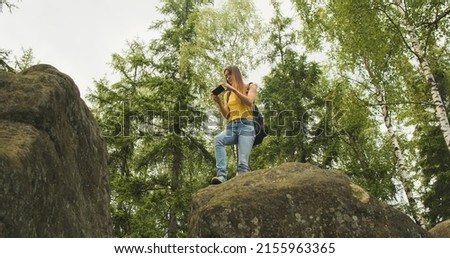 young girl climbed a hill and takes pictures in the forest on her smartphone, standing on a stone. Green trees on the background, bottom view