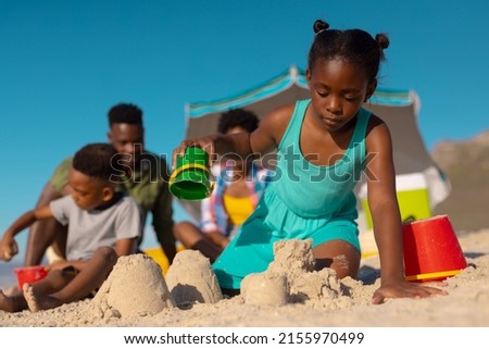Low angle view of african american girl making sand castle against sky with family in background. copy space, unaltered, beach, childhood, family, togetherness, lifestyle, enjoyment, holiday.